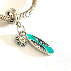 Może przedstawiać: Srebrna bransoletka z zawieszką w kształcie turkusowej deski surfingowej i zawieszką w kształcie słońca z żółtym środkiem. Zawieszka deski surfingowej ma srebrną obwódkę i czarne detale. Zawieszki są przymocowane do srebrnej bransoletki.