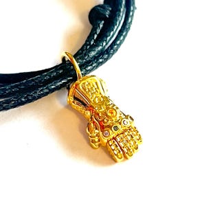 Könnte beinhalten: Ein Armband aus geflochtenem schwarzem Band mit einem goldfarbenen Infinity-Handschuh-Anhänger. Der Handschuh ist mit detaillierten Gravuren und bunten Edelsteinen verziert.