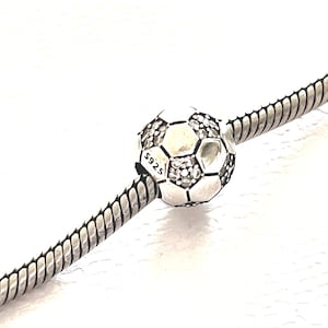 Puede incluir: Un charm plateado con forma de balón de fútbol, con un patrón de paneles hexagonales y pentagonales. El charm está adornado con pequeñas gemas transparentes. El texto "S925" es visible. El charm está ensartado en una cadena plateada.