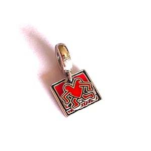 Peut inclure: Un charm en argent avec un motif rouge et noir. Le charm représente un cœur rouge et une figure stylisée sur fond rouge. Le charm est de forme carrée et possède une boucle argentée pour l'attacher à un bracelet ou un collier.