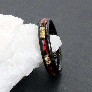 Puede incluir: Un anillo negro con detalles rojos y dorados. El anillo presenta una banda de motas rojas y doradas incrustadas en un material negro. El anillo se muestra sobre una superficie blanca y texturizada sobre un fondo oscuro.