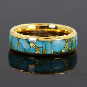 Può includere: Un anello color oro con una fascia di pietre color turchese e dettagli color oro. L'anello è posto su uno sfondo scuro, con un riflesso dell'anello visibile sotto.