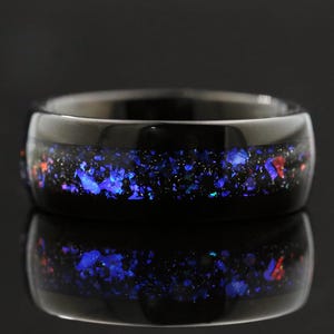 Könnte beinhalten: Ein schwarzer Ring mit einem Band aus funkelnden blauen, roten und violetten Fragmenten. Der Ring ist vor einem dunklen Hintergrund mit einer reflektierenden Oberfläche darunter platziert. Das Design des Rings ähnelt einer Galaxie.