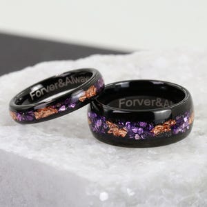 Può includere: Due anelli neri con la scritta "Forver & Always". Gli anelli presentano una fascia di accenti viola e color rame. Un anello è più sottile dell'altro. Gli anelli sono esposti su una superficie bianca.