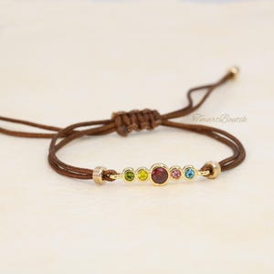 Puede incluir: Un brazalete de cordón marrón con una barra dorada y siete piedras preciosas de colores. Las piedras preciosas son verdes, amarillas, rojas, rosas, azules y transparentes.