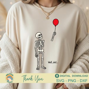May include: A cream-colored t-shirt featuring a black line art graphic of a skeleton looking up at a detached skeletal arm holding a single red balloon. The text "Well, shit..." is printed in black ink next to the skeleton on the front of the shirt.