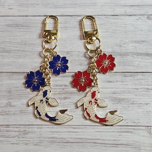 May include: Two gold-toned keychains with koi fish charms. Each keychain features a fish charm with white and colored accents, and a flower charm in either red or blue. The keychains have gold-colored clasps.