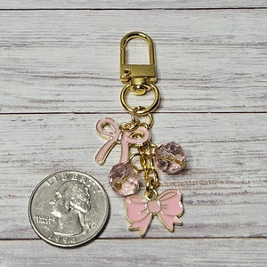 May include: A gold-colored keychain with pink bow charms and faceted pink beads. The keychain includes a lobster clasp and a quarter for size comparison. The bows and beads are suspended from a gold chain.