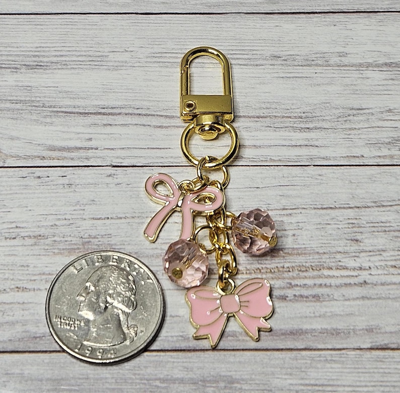 May include: A gold-colored keychain with pink bow charms and faceted pink beads. The keychain includes a lobster clasp and a quarter for size comparison. The bows and beads are suspended from a gold chain.