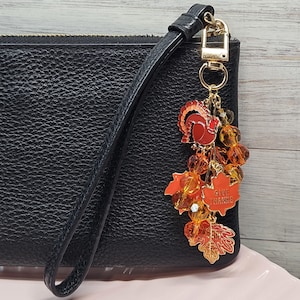 May include: A black textured wristlet featuring a gold-tone metal keychain charm. The charm includes a red turkey, orange maple leaves, amber-coloured faceted beads, and a leaf-shaped pendant with the engraved text GIVE THANKS in gold lettering.