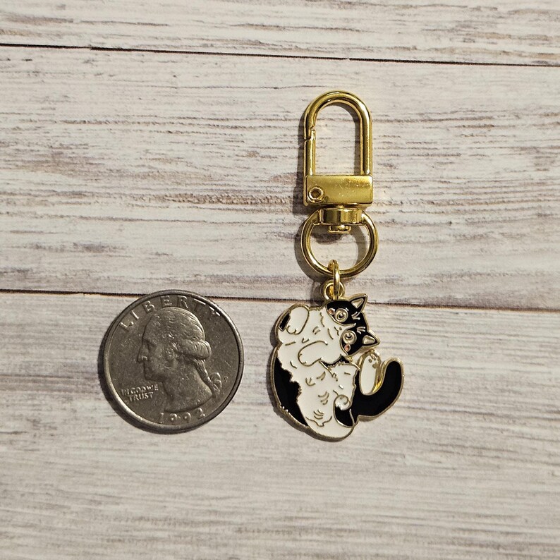 May include: A gold-colored metal keychain with a black and white cat charm. The charm is a cartoon cat design with a gold-colored clasp and ring. A US quarter is next to the keychain.