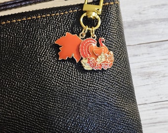 May include: A gold-toned charm with a red maple leaf and a turkey surrounded by fall leaves. The charm is attached to a black leather pouch with a gold clasp. The turkey charm is orange, red, and yellow.