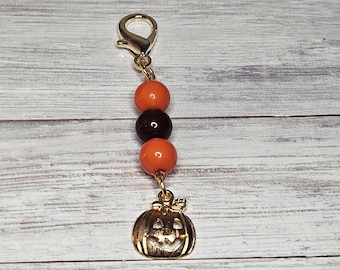 May include: A gold-colored Halloween-themed charm with a lobster clasp. The charm features three beads in orange and brown, and a gold-colored pumpkin pendant with a carved face. The charm is designed for use as a keychain or zipper pull.