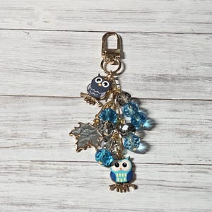 May include: A gold-toned metal keychain featuring two owl charms, one dark gray and one light blue, alongside a silver-toned maple leaf charm. The design includes multiple faceted beads in shades of translucent blue and dark gray, all attached to a lobster clasp.