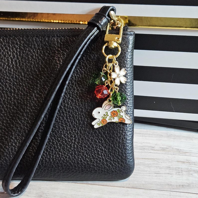 May include: Black leather wristlet with a gold-tone clasp and a charm. The charm features a white bunny with orange floral accents, a white flower, and green and red beads. The wristlet is on a light wood surface.