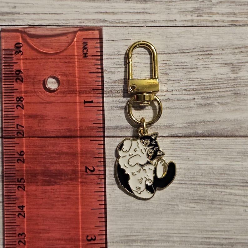 May include: A gold-colored metal keychain with a black and white cat charm. The cat charm is approximately 1.5 inches long and features a playful design. The keychain has a lobster clasp for easy attachment.