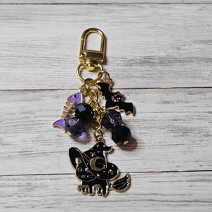 Op de afbeelding: Een goudkleurige metalen sleutelhanger met een karabijnsluiting en een gouden ketting. De bedels bestaan uit een paars met zilverkleurige vissengraat, een zwarte vleermuis en een zwarte kat met een heksenhoed en een halve maan. Donkerpaarse kralen.