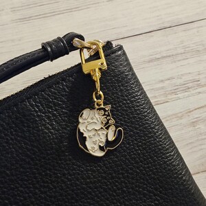 May include: A gold-colored cat charm with black and white enamel detailing hangs from a black leather wristlet. The charm features a stylized cat design. The wristlet has a gold zipper and a black leather strap.