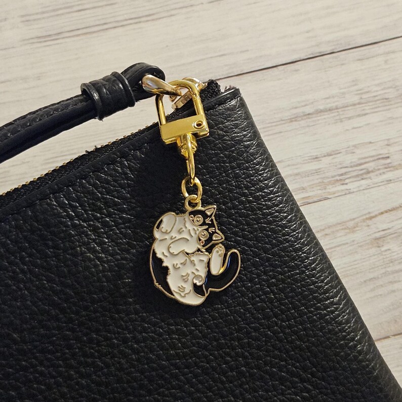 May include: A gold-colored cat charm with black and white enamel detailing hangs from a black leather wristlet. The charm features a stylized cat design. The wristlet has a gold zipper and a black leather strap.