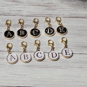 May include: Twelve gold-toned metal letter charms with black and white enamel. The charms feature the letters A, B, C, D, and E. Each charm has a small lobster clasp.