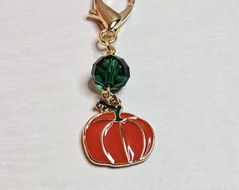 May include: A gold-colored lobster clasp charm with a red pumpkin pendant. The pumpkin has a gold outline and green stem. A faceted green bead is between the clasp and the pumpkin.