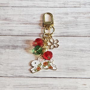 May include: A gold-colored keychain with a white rabbit charm decorated with orange flowers. The keychain also features red and green beaded accents, a white flower charm, and a gold clasp.