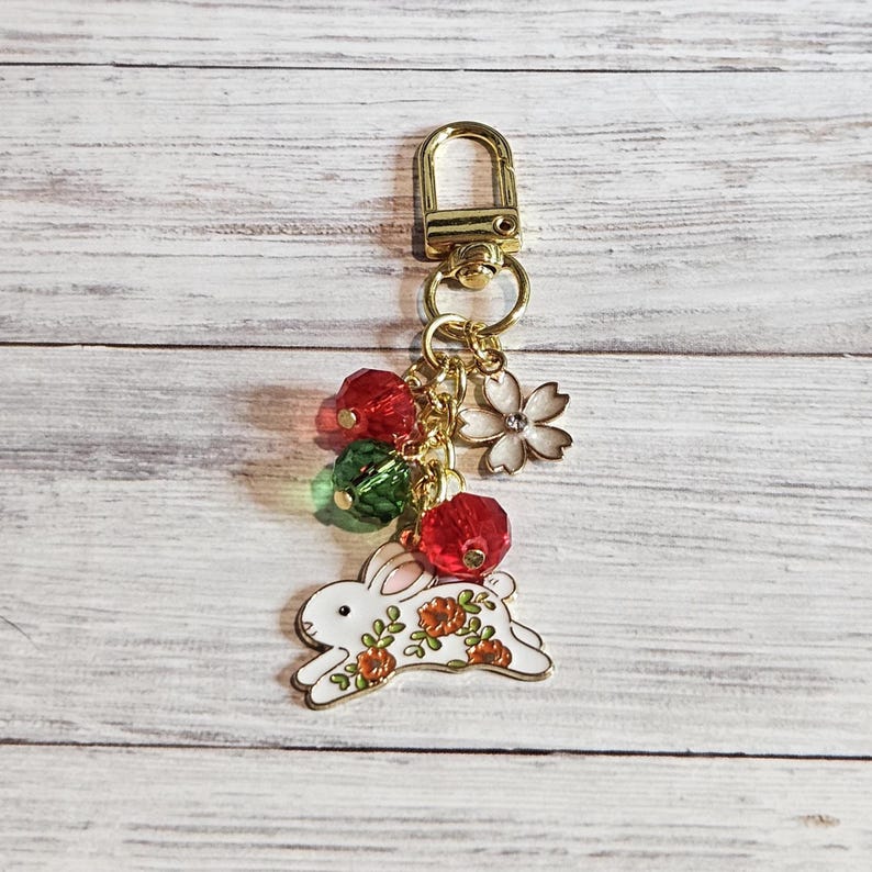 May include: A gold-colored keychain with a white rabbit charm decorated with orange flowers. The keychain also features red and green beaded accents, a white flower charm, and a gold clasp.