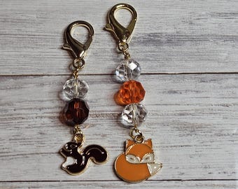 May include: Two charm keychains with gold-coloured clasps. One features a brown squirrel charm, the other an orange fox charm. Both have clear and brown or orange beads. The charms are attached to the clasps with gold-coloured chains.