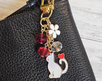 May include: A gold-toned keychain charm featuring a white cat with a red heart, white flower, red and clear crystal beads. The charm is attached to a black leather wallet with a gold zipper.