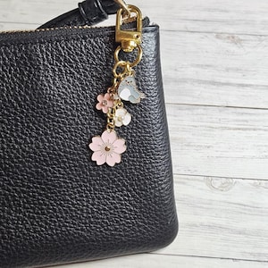 May include: A gold-toned keychain charm with a black leather bag. The charm features a gray and white cartoon seal, pink and white flowers, and a pink cherry blossom. The bag has a black strap and a gold-toned clasp.