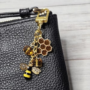 May include: A gold-colored charm with a bee and honeycomb design, attached to a black leather item. The bee has yellow and black stripes, with crystal accents on the wings. The honeycomb is a hexagonal shape.