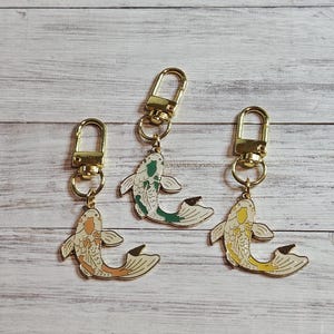 May include: Three keychains with gold-colored hardware and koi fish charms. The koi charms feature different color schemes: orange and white, green and white, and yellow and white. The keychains are displayed on a light wood surface.