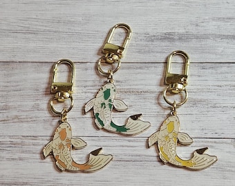 May include: Three keyrings with gold-coloured hardware and koi fish charms. The koi charms feature different colour schemes: orange and white, green and white, and yellow and white. The keyrings are displayed on a light wood surface.