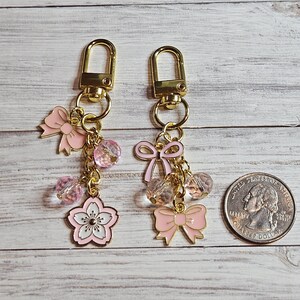 May include: Two keychains with gold-colored clasps and chains. Each keychain features pink bows, clear and pink beads, and a pink and white cherry blossom charm. A US quarter is included for size reference.