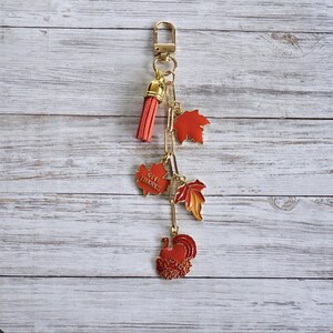 Op de afbeelding: Een goudkleurige sleutelhanger met een rode kwast en een reeks bedels. De bedels omvatten rode esdoornbladeren, een kalkoen en een bedel met de tekst "GIVE THANKS". De sleutelhanger heeft een herfst- of Thanksgiving-thema.