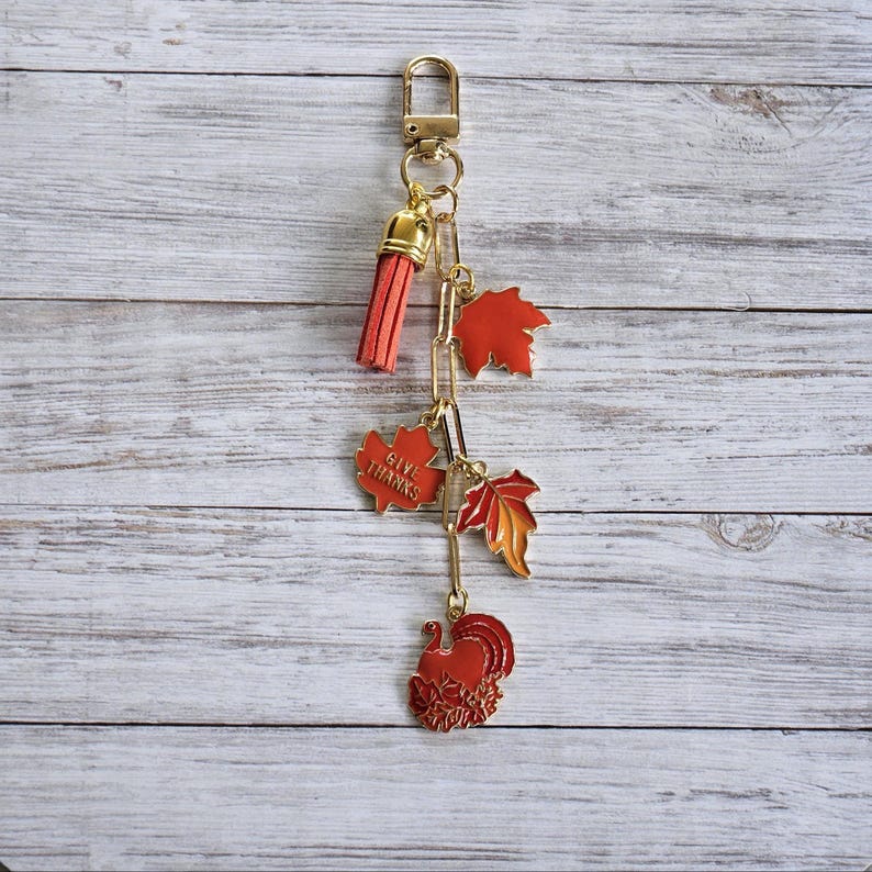 Op de afbeelding: Een goudkleurige sleutelhanger met een rode kwast en een reeks bedels. De bedels omvatten rode esdoornbladeren, een kalkoen en een bedel met de tekst "GIVE THANKS". De sleutelhanger heeft een herfst- of Thanksgiving-thema.