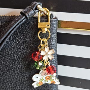 May include: A gold-toned keychain charm with a white bunny, red and green faceted beads, and a white flower charm. The bunny has orange floral accents. The charm is attached to a black leather wristlet.