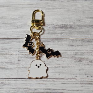 Op de afbeelding: Een goudkleurige metalen sleutelhanger met een karabijnsluiting en een korte ketting. Aan de ketting hangen drie emaille bedels: twee zwarte vleermuizen met lachende gezichten en een wit spookje met een hartvormig mondje en twee zwarte oogjes.