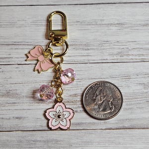 May include: A gold-colored keychain with a lobster clasp, featuring a pink bow, two pink crystal beads, and a pink and white cherry blossom charm. A U.S. quarter is next to the keychain for size comparison.