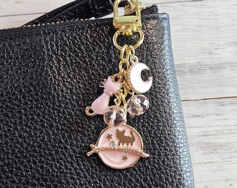 May include: A gold-toned keychain charm with a pink cat, a crescent moon, and a planet design. The charm is attached to a black leather wallet. The charm features a cat silhouette, stars, and clear crystal beads.