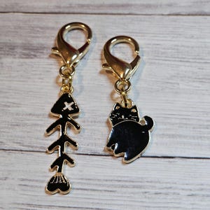 May include: Two gold-toned metal charms with lobster clasps. One charm features a black fishbone with a white cross on the head. The other charm features a black cat with white eyes.