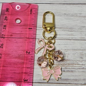 May include: A gold-colored keychain with pink bow charms and faceted pink beads. The keychain includes a lobster clasp and a ruler showing inches.