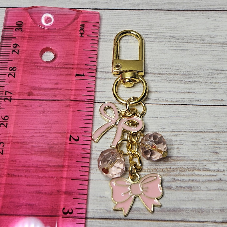 May include: A gold-colored keychain with pink bow charms and faceted pink beads. The keychain includes a lobster clasp and a ruler showing inches.