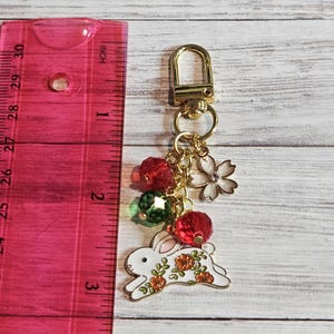 May include: Gold-tone keychain with a white rabbit charm decorated with orange flowers. The keychain also features red and green beads, a white flower charm, and a lobster clasp. The keychain measures approximately 3 inches in length.