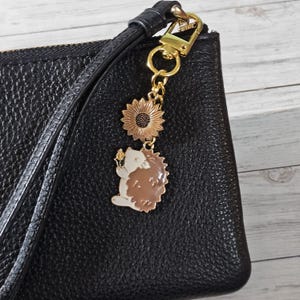 May include: A black textured leather wristlet featuring a gold-tone metal keychain attachment. The keychain includes a sunflower charm and an enamel hedgehog charm in shades of white and brown, suspended from a gold-tone lobster clasp.