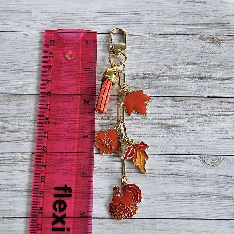 Op de afbeelding: Een goudkleurige sleutelhanger met een kwastje en Thanksgiving-thema bedels. De bedels omvatten oranje en rode herfstbladeren, een kalkoen en een bedel met de tekst "GIVE THANKS". De sleutelhanger is ongeveer 13 cm lang.