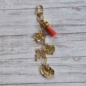 Op de afbeelding: Goudkleurige sleutelhanger met een karabijnhaak, ketting en bedels. De bedels omvatten bladvormen en een kalkoen. Een koraalkleurige kwast hangt van bovenaf. De achtergrond is een lichte houtnerf.