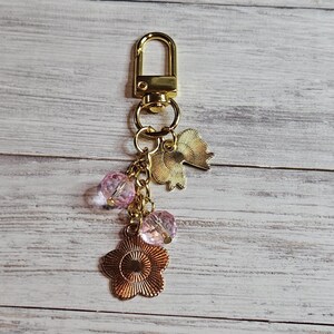 May include: Gold-toned keychain with a lobster clasp, featuring a flower charm, two pink crystal beads, and a gold fan-shaped charm. The keychain is designed with a decorative aesthetic.
