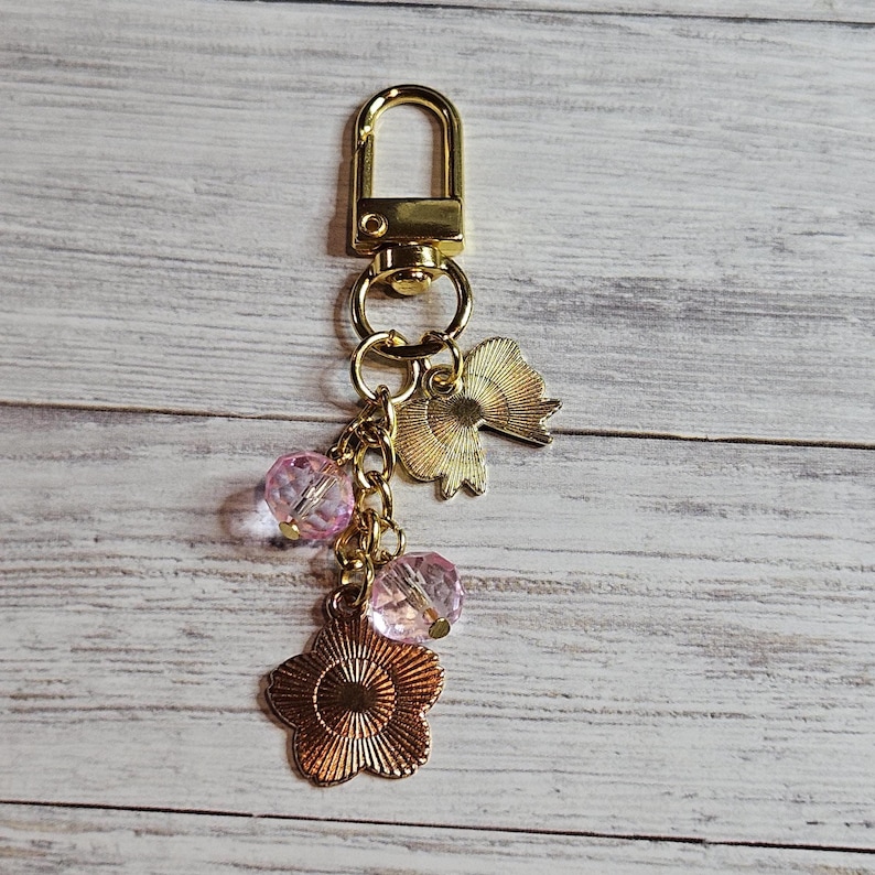 May include: Gold-toned keychain with a lobster clasp, featuring a flower charm, two pink crystal beads, and a gold fan-shaped charm. The keychain is designed with a decorative aesthetic.
