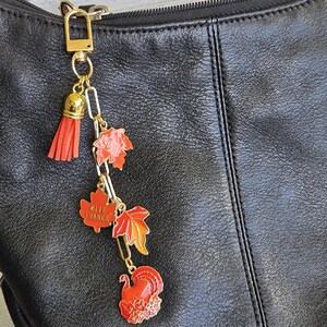 Op de afbeelding: Een goudkleurige tashanger met een rode kwast en herfstcharms. De bedels tonen esdoornbladeren, &eacute;&eacute;n met de tekst "GIVE THANKS", en een kalkoen. De hanger is bevestigd aan een zwarte leren tas.